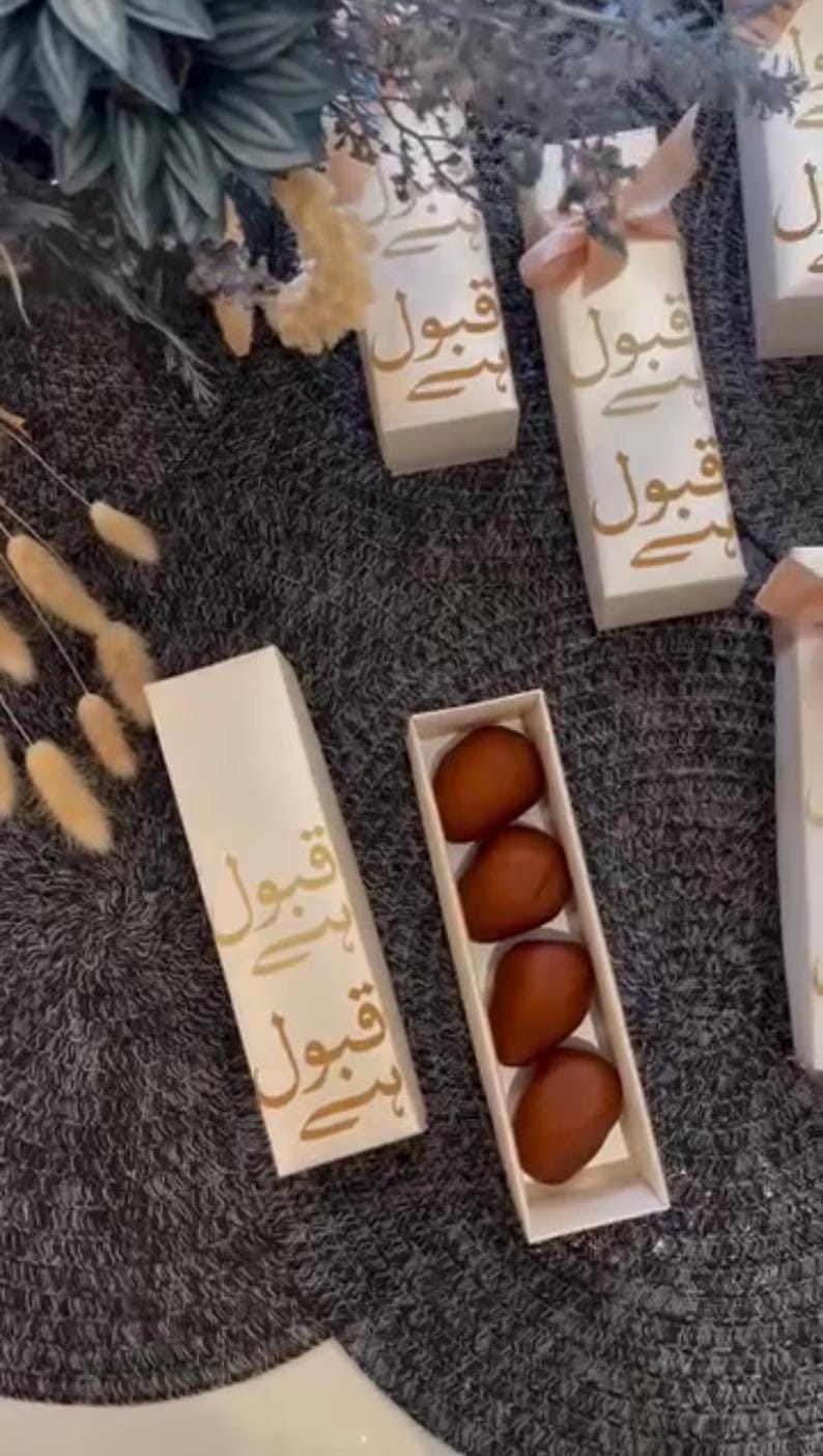 Pu&ograve; includere: Diverse scatole regalo rettangolari bianche con scritta araba dorata sono disposte su una superficie grigia. Una scatola aperta rivela tre dolcetti marroni di forma ovale.