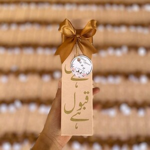 Könnte beinhalten: Eine beige Geschenkbox mit goldener Schrift und einer goldenen Schleife. Die Box hat goldene Schrift in einer Fremdsprache. Ein rundes Etikett mit floralen Akzenten und Text hängt an der Schleife. Die Box wird in einer Hand gehalten.