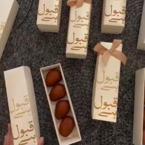 Pu&ograve; includere: Scatole regalo color crema con testo dorato e fiocco color pesca. Sono disposte su una superficie grigia. Una scatola rettangolare contiene datteri marroni ovali. Il testo &egrave; in lingua straniera.
