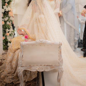 May include: A bride in a cream wedding dress with gold embroidery and a long, sheer veil with gold sequins. She sits on a vintage white chair with carvings, holding a pink rose. The scene suggests a wedding ceremony.