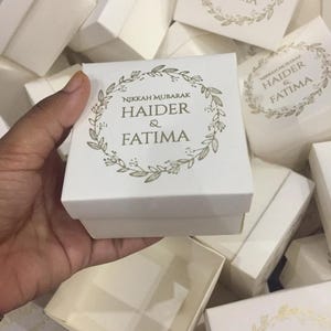 Puede incluir: Cajas de regalo cuadradas blancas con texto dorado y diseño de corona floral. El texto en la caja dice "Nikkah Mubarak Haider & Fatima". Las cajas están apiladas, con una caja abierta, revelando el interior.