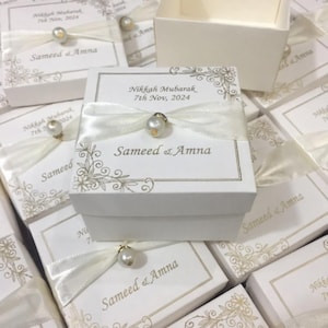 May include: White gift boxes tied with ivory ribbons and adorned with pearl embellishments. The boxes feature floral patterns and the text "Nikkah Mubarak, 7th Nov, 2024" and "Sameed & Amna".