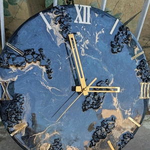 Puede incluir: Un reloj de pared redondo con una esfera azul oscuro y dorada veteada. El reloj presenta manecillas doradas y números romanos. Acentos negros texturizados se colocan alrededor de la esfera.