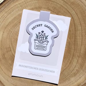Könnte beinhalten: Rechteckiger magnetischer Lesezeichen mit einer Illustration einer Schreibmaschine und Blumen auf grauem Hintergrund. Der Text "SECRET GARDEN" befindet sich darüber. Das Lesezeichen trägt auch den Text "MAGNETISCHES LESEZEICHEN" und "@FluffnCraft".