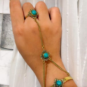 Puede incluir: Una pulsera de mano dorada con tres cabujones redondos de color turquesa engastados en monturas de metal con filigrana. La pieza consta de un anillo conectado por una cadena fina a un elemento decorativo central y a una pulsera de muñeca.