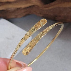 May include: A gold-toned bypass bracelet featuring a smooth, thin wire band that transitions into two flat, overlapping ends with a textured, hammered finish. The metal surface reflects light, highlighting the irregular indentations across the ends.