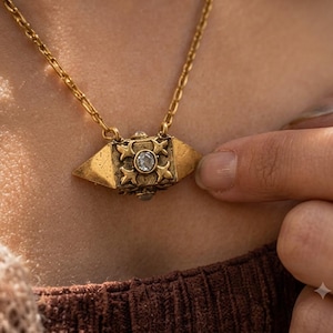 Puede incluir: Un collar de tono dorado con un colgante geométrico puntiagudo que presenta una piedra clara central engastada en un marco de metal ornamentado en forma de cruz. El colgante cuelga de una delicada cadena dorada con detalles texturizados.