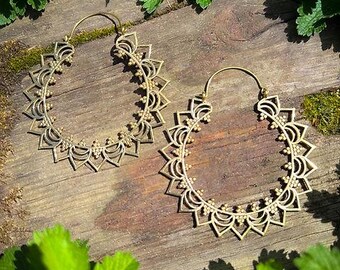 May include: Two gold-colored hoop earrings with a sunburst design. The earrings are intricately detailed with a series of pointed shapes and small circular accents. The earrings are displayed on a wooden surface with green foliage.