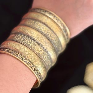 May include: A stack of antique gold-colored metal bracelets with intricate floral and geometric patterns. The bracelets are worn on a wrist, showcasing the detailed designs and textures. The bracelets have a vintage, bohemian aesthetic.