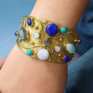 May include: A wide, ornate gold-toned cuff bracelet. The bracelet is adorned with various gemstones, including blue lapis lazuli, turquoise, and moonstone. The bracelet is approximately 2 inches wide.