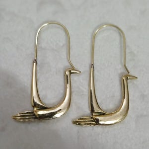 May include: A pair of gold-colored bird-shaped earrings. The earrings feature a stylized swan design with a curved body and a long, thin neck. The earrings are displayed on a light-colored surface.