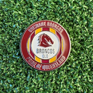 May include: A circular metal medallion displaying the Brisbane Broncos logo with a horse head silhouette. The text reads BRISBANE BRONCOS, BRONCOS QLD, and PRIDE OF QUEENSLAND. The design features maroon, gold, and white enamel colour detailing.