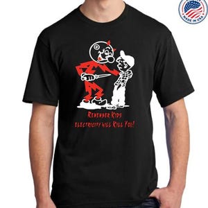 May include: Black t-shirt featuring a cartoon illustration of a red figure with a knife confronting a white figure. The text "Remember Kids Electricity Will Kill You!" is printed below the figures. A "Proudly Made in USA" emblem is visible.
