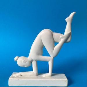May include: A white sculpture of a person in a yoga pose, balancing on hands with one leg extended upwards. The figure is on a rectangular base. The background is a solid blue.