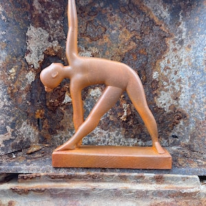 Può includere: Una scultura di posa yoga color bronzo. La figura è in una posa eretta con un braccio alzato e l'altro che raggiunge il piede. La scultura è montata su una base rettangolare.