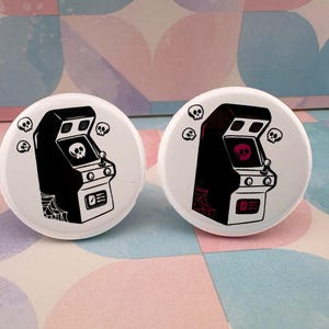 May include: Two white buttons with black and red arcade machine designs. Each button features a black arcade machine with a skull on the screen. One skull is black, the other is red. Small skulls surround the arcade machines.
