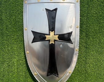 May include: A silver-colored shield with a black cross design and a gold cross in the center. The shield has a rounded bottom and is edged with small gold rivets. The shield is displayed on a green surface.