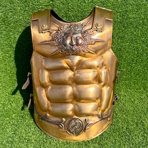 May include: A gold-coloured breastplate with a sculpted design. The chest armour features a detailed depiction of a face with radiating lines and a belt buckle with a circular emblem. The breastplate has a sculpted, muscular appearance.