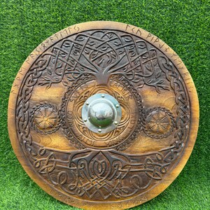 May include: A circular wooden shield with detailed carvings. The design includes a central tree motif, knotwork, and runic inscriptions. A silver metal boss is at the centre. The shield has a warm brown stain.