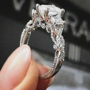 May include: A silver engagement ring with a large, square-cut center stone and smaller side stones. The band features intricate detailing and is encrusted with smaller accent diamonds. The ring is held up by a finger.
