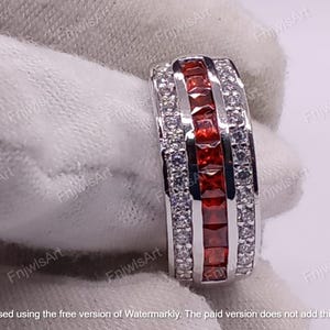 May include: A silver ring with a rectangular design. The ring features a row of square, red gemstones flanked by rows of smaller, clear gemstones. The ring is set against a white background.