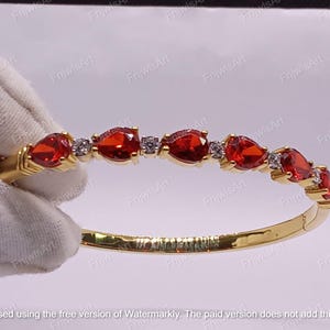 Puede incluir: Una pulsera dorada adornada con una serie de gemas rojas vibrantes en forma de lágrima. La pulsera está acentuada con pequeñas piedras transparentes y tiene un cierre de bisagra. Una mano enguantada de blanco sostiene la pulsera.