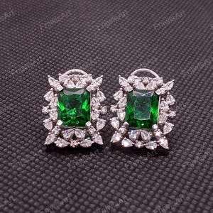 Puede incluir: Un par de pendientes de botón con una gran piedra preciosa verde esmeralda en el centro, rodeada de piedras de acento más pequeñas, claras y brillantes. Los pendientes están engarzados en un metal plateado.