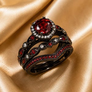 May include: A black metal ring set featuring a large round red center stone surrounded by a halo of small white stones. The bands are decorated with alternating red and white stones in a filigree pattern, with a V-shaped band included.