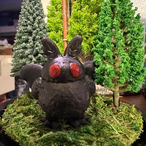 K&ouml;nnte beinhalten: Eine schwarze, 3D-gedruckte Fledermausfigur mit roten Augen, die auf einem Bett aus gr&uuml;nem Moos sitzt. Die Fledermaus ist von kleinen immergr&uuml;nen B&auml;umen umgeben.