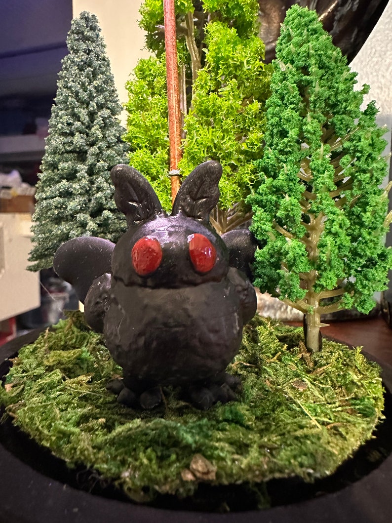 K&ouml;nnte beinhalten: Eine schwarze, 3D-gedruckte Fledermausfigur mit roten Augen, die auf einem Bett aus gr&uuml;nem Moos sitzt. Die Fledermaus ist von kleinen immergr&uuml;nen B&auml;umen umgeben.