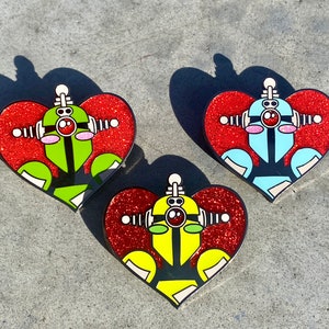 以下が含まれることがあります： キラキラ光るハート型のピンが3つ。それぞれのピンには、緑、青、黄色の異なる色のボディを持つロボットが描かれています。ロボットは赤い目を持ち、アンテナがあります。