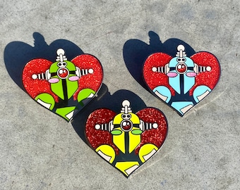 Op de afbeelding: Drie hartvormige pinnen met glitter. Elke pin heeft een robot met een andere kleur lichaam: groen, blauw en geel. De robots hebben rode ogen en antennes.