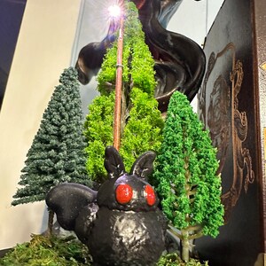 K&ouml;nnte beinhalten: Eine schwarze, 3D-gedruckte, mottenartige Kreatur mit roten Augen sitzt in einer Miniatur-Waldszene. Die Szene enth&auml;lt drei kleine, gr&uuml;ne, k&uuml;nstliche B&auml;ume und einen braunen Stock mit einem Licht an der Spitze. Die Szene ist auf einem Bett aus gr&uuml;nem Moos platziert.