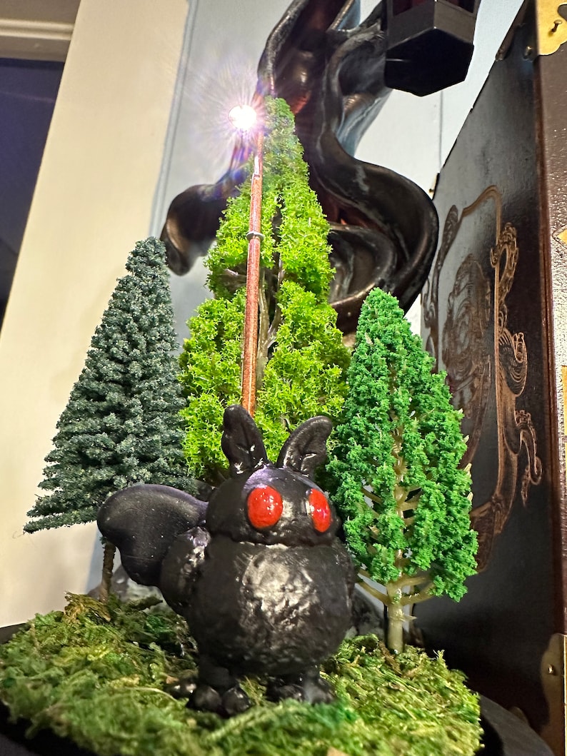 K&ouml;nnte beinhalten: Eine schwarze, 3D-gedruckte, mottenartige Kreatur mit roten Augen sitzt in einer Miniatur-Waldszene. Die Szene enth&auml;lt drei kleine, gr&uuml;ne, k&uuml;nstliche B&auml;ume und einen braunen Stock mit einem Licht an der Spitze. Die Szene ist auf einem Bett aus gr&uuml;nem Moos platziert.