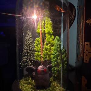 K&ouml;nnte beinhalten: Ein Glas-Terrarium in Kuppelform mit einem kleinen, dunkelgrauen, fledermausartigen Wesen mit roten Augen. Das Wesen ist umgeben von gr&uuml;nem Moos und kleinen, gr&uuml;nen, k&uuml;nstlichen B&auml;umen. Ein Licht scheint durch die Oberseite der Kuppel.