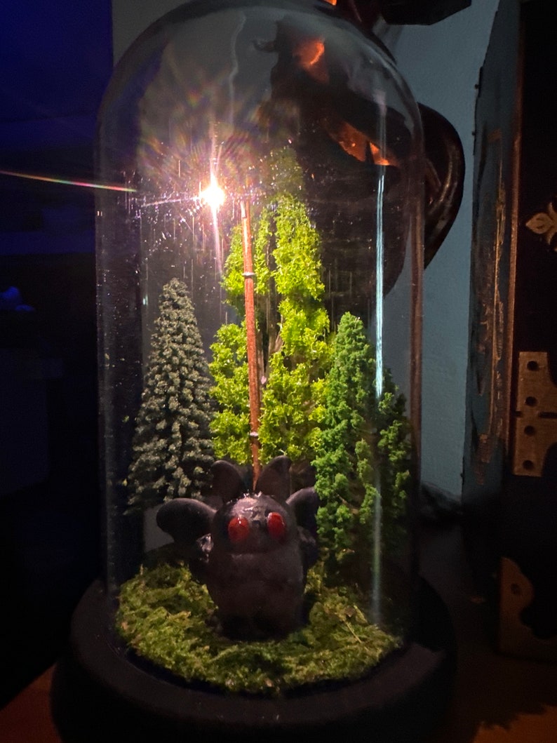 K&ouml;nnte beinhalten: Ein Glas-Terrarium in Kuppelform mit einem kleinen, dunkelgrauen, fledermausartigen Wesen mit roten Augen. Das Wesen ist umgeben von gr&uuml;nem Moos und kleinen, gr&uuml;nen, k&uuml;nstlichen B&auml;umen. Ein Licht scheint durch die Oberseite der Kuppel.