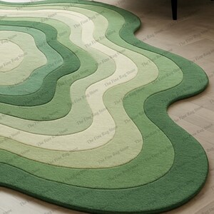 Puede incluir: Una alfombra verde con bordes ondulados y un dise&ntilde;o en capas. La alfombra presenta tonos de verde, de claro a oscuro, creando un efecto degradado. The Fine Rug Store est&aacute; impreso en la alfombra.