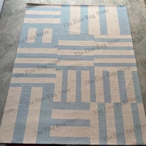 Puede incluir: Una alfombra rectangular con un patrón geométrico de rayas y bloques en azul claro y beige. La alfombra tiene una textura tejida y es adecuada para una sala de estar o un dormitorio. The Fine Rug Store está impreso en la alfombra.