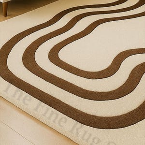 K&ouml;nnte beinhalten: Cremefarbener Teppich mit einem sich wiederholenden Muster aus wellenf&ouml;rmigen, braunen Streifen. Der Teppich liegt auf einem Holzboden, im Hintergrund ist ein beiges Sofa zu sehen. Das Design des Teppichs verleiht dem Raum eine moderne Note.