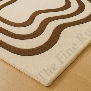 K&ouml;nnte beinhalten: Cremefarbener Teppich mit braunem, wellenf&ouml;rmigem Muster. Das Design des Teppichs weist drei geschwungene Linien auf, die einen flie&szlig;enden, organischen Look erzeugen. Der Teppich liegt auf einem hellen Holzboden, wobei die Ecke des Teppichs sichtbar ist.