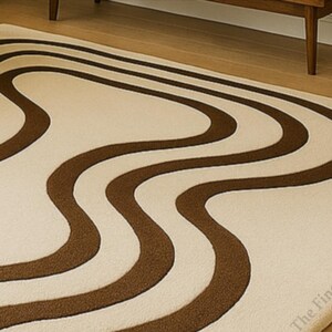K&ouml;nnte beinhalten: Ein cremefarbener Teppich mit einem sich wiederholenden braunen Wellenmuster. Der Teppich hat eine weiche Textur und ist rechteckig. Das Design erinnert an einen Retro-Stil.