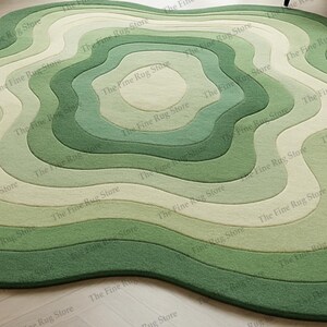 Puede incluir: Una alfombra verde con bordes ondulados y un dise&ntilde;o en capas. La alfombra presenta formas conc&eacute;ntricas y org&aacute;nicas en diferentes tonos de verde y blanco roto, creando un revestimiento de suelo texturizado. The Fine Rug Store est&aacute; impreso en la alfombra.