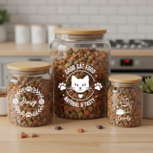 Puede incluir: Tres tarros de vidrio con tapas de madera llenos de comida para gatos. Los tarros tienen etiquetas blancas con diseños y texto con temática de gatos, incluyendo "Good Cat Food", "Daisy" y "treats". Los tarros están sobre una superficie de madera.
