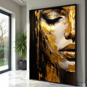 May include: Large framed art print depicting a close-up of a woman's face. The artwork uses gold and black paint to create an abstract portrait. The woman's lips and parts of her face are painted gold, with long black eyelashes.