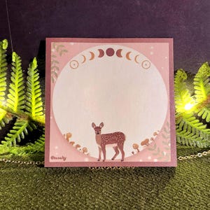 May include: A square, pink sticky note with a deer illustration. The design includes a white circle for writing, moon phases, mushrooms, and foliage. The text "@tasalty" is in the lower left corner.