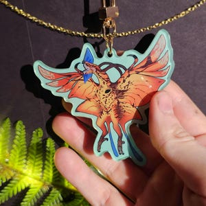May include: A colorful acrylic charm featuring a stylized orange and yellow dragon with outstretched wings. The charm has a light blue border and is attached to a gold-colored chain and clasp. The dragon has blue accents and a detailed design.