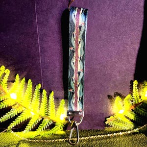 May include: A patterned wristlet key fob with a silver-tone clasp. The design features a repeating pattern of birds on a tree branch with a wavy background in shades of blue, green, and purple. The wristlet is made of a durable material.