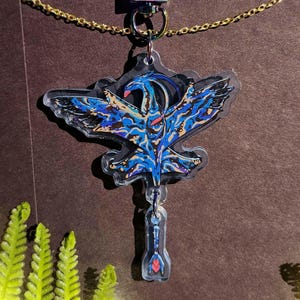 May include: A clear acrylic pendant with a blue and black winged creature design, red beak and accents. The pendant hangs from a gold-coloured chain. The design is detailed with blue, black, and yellow patterns. The pendant has a small, clear, arrow-shaped charm attached.
