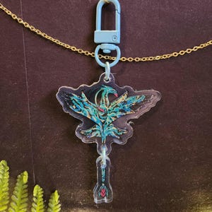 以下が含まれることがあります： 水色のカラビナが付いたアクリル製キーホルダー。黒とターコイズブルーの模様が入った翼を持つ鳥のような生き物が描かれています。下部には矢の形をした小さなチャームがぶら下がっています。