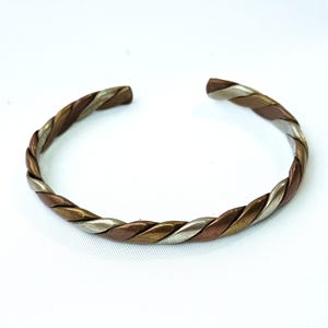 May include: A braided cuff bracelet in silver, copper, and gold. The open-ended bracelet features a twisted design, with the three metal colours interwoven. The bracelet is displayed against a white backdrop.